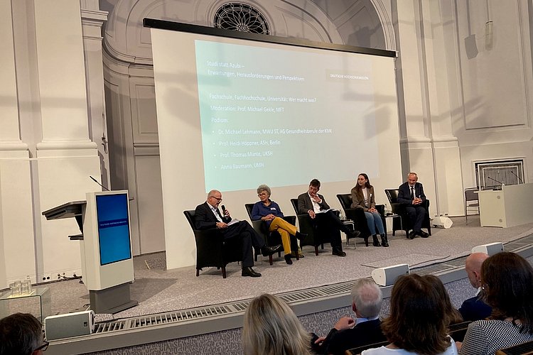 The photo shows people on a podium, including Prof. Dr. Heidi Höppner as a panel member.